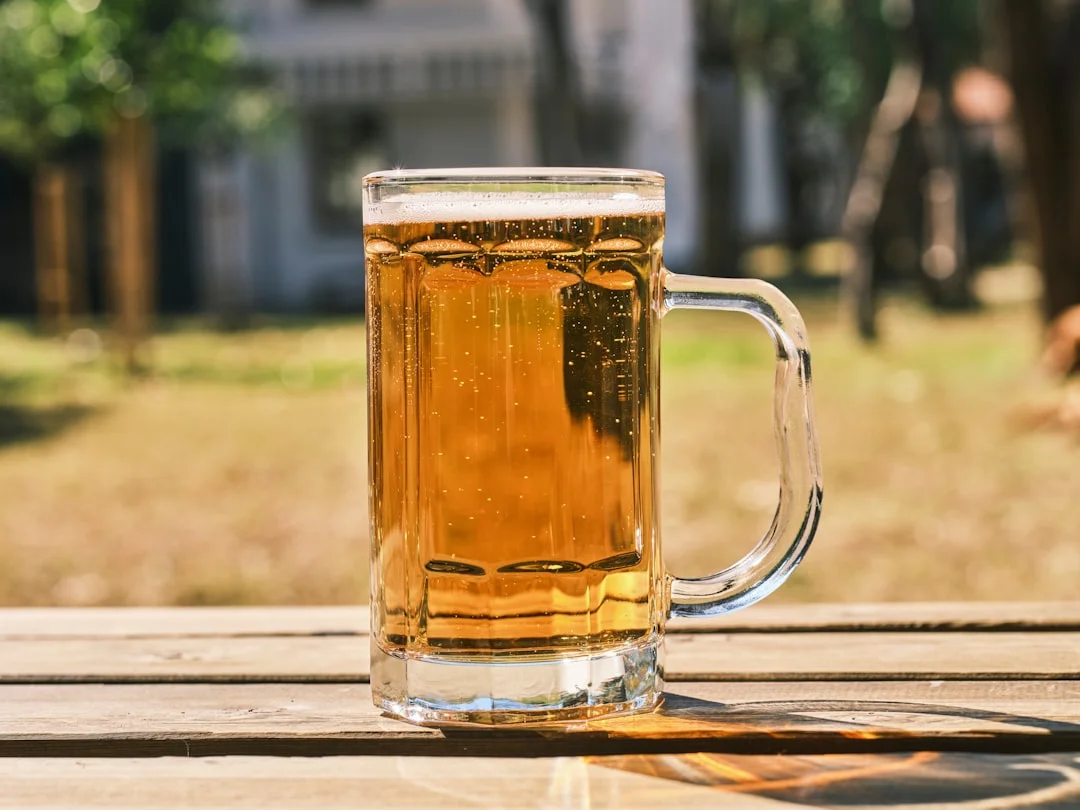 A stainless steel all-in-one electric brewing system on a garage workbench, surrounded by a grain mill, hop pellets in open bags, and a glass of golden homebrew